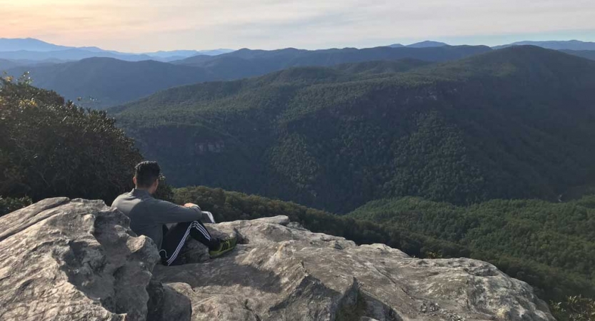 A person sits on a rocky overlook, looking out over the vast forested mountain area below. 